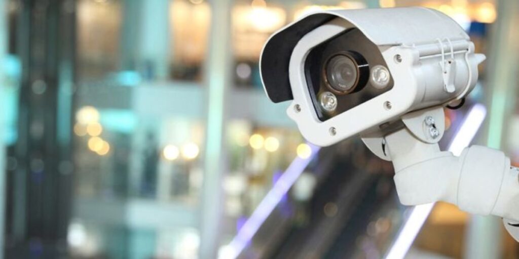 Surveillance camera in focus with blurred mall background and escalator lights in the distance.