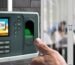 Person using a biometric fingerprint scanner for attendance outside a glass-walled office meeting room.