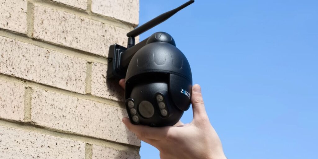 Person installing a black wireless CCTV security camera on a brick wall under clear blue sky.