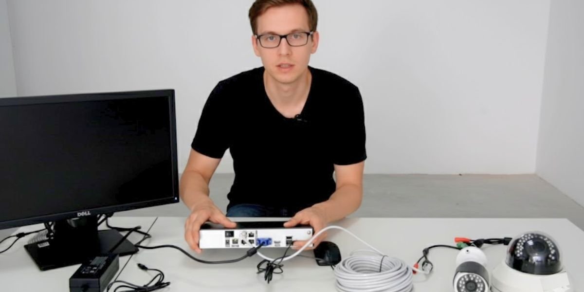 A man with glasses and a black shirt sits at a desk with a monitor, DVR, cables, and two security cameras.