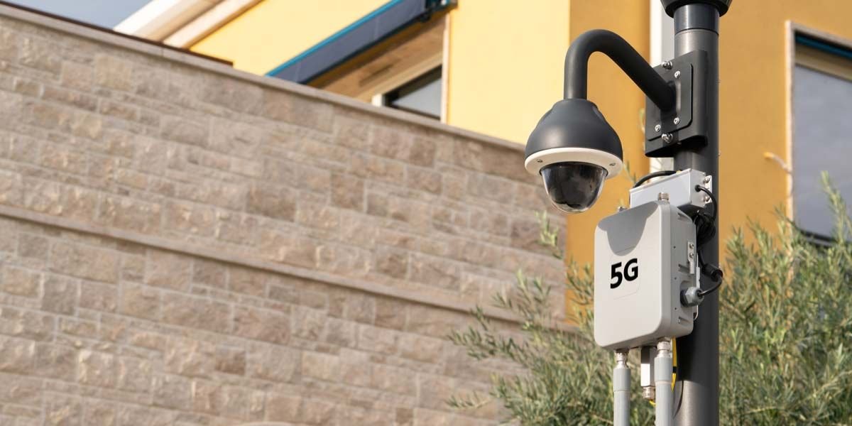 A black dome 5G CCTV camera is mounted on a pole with a box labeled 5G in front of a building