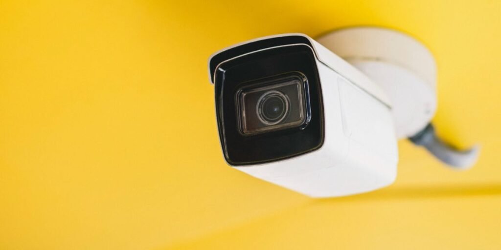 A white, bullet-style security camera mounted on a bright yellow wall or ceiling, looking downward