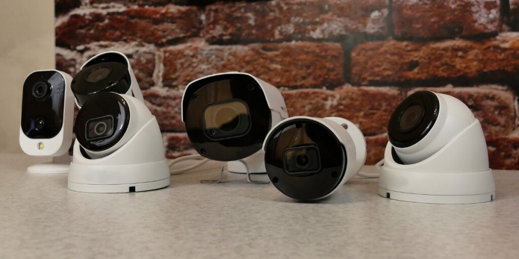 Various white dome and bullet security cameras of different sizes arranged on a table against a red brick wall.
