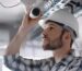 A technician in a hard hat and plaid shirt installs a white security camera onto a ceiling with exposed wiring.