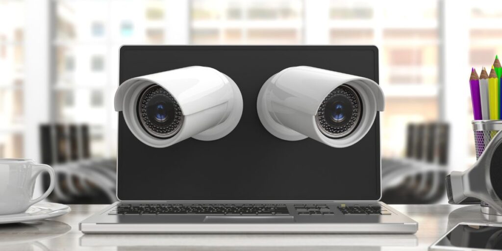 Two white security cameras appearing to emerge from a laptop screen on a desk with a coffee cup and pencils.
