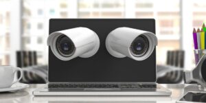 Two white security cameras appearing to emerge from a laptop screen on a desk with a coffee cup and pencils.