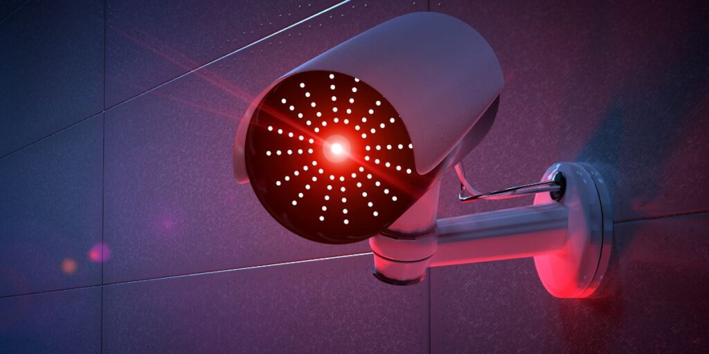 White bullet security camera with bright red infrared lights mounted on a dark wall in a dim, purple-lit setting.