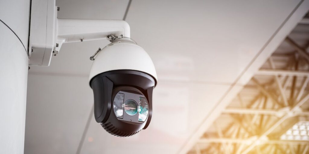 A white and black PTZ dome security camera mounted to a ceiling, monitoring a large indoor facility with bright light.