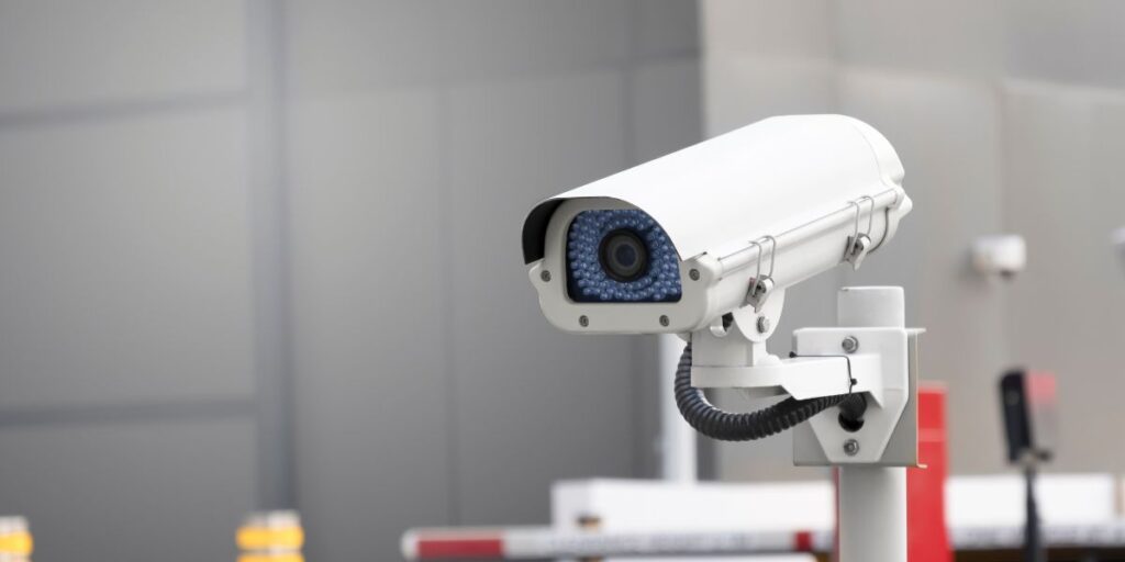 A white outdoor bullet CCTV security camera mounted on a pole, monitoring a modern industrial or commercial area.
