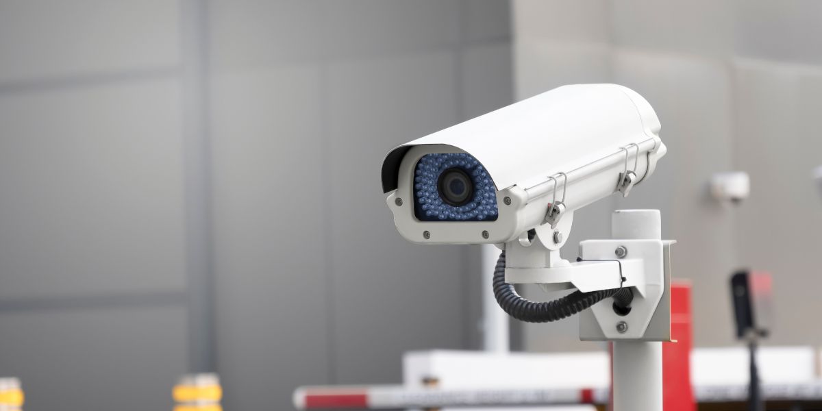 A white outdoor bullet CCTV security camera mounted on a pole, monitoring a modern industrial or commercial area.