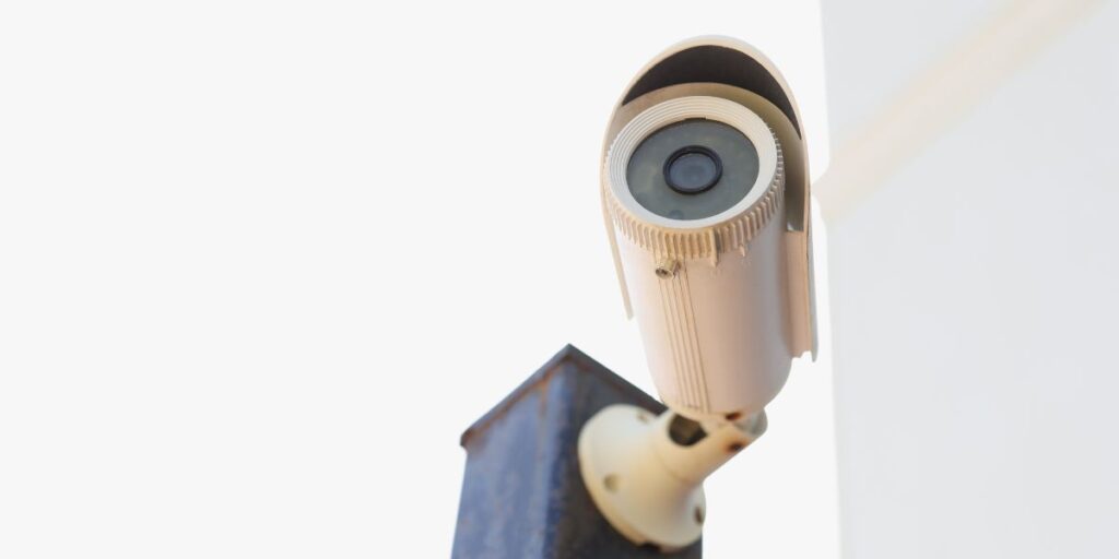A low-angle shot of a white bullet CCTV security camera mounted on a metal post against a bright, clear sky.